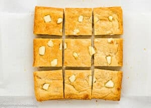 A batch of blondies cut into nine squares, each topped with pieces of white chocolate, placed on white parchment paper.