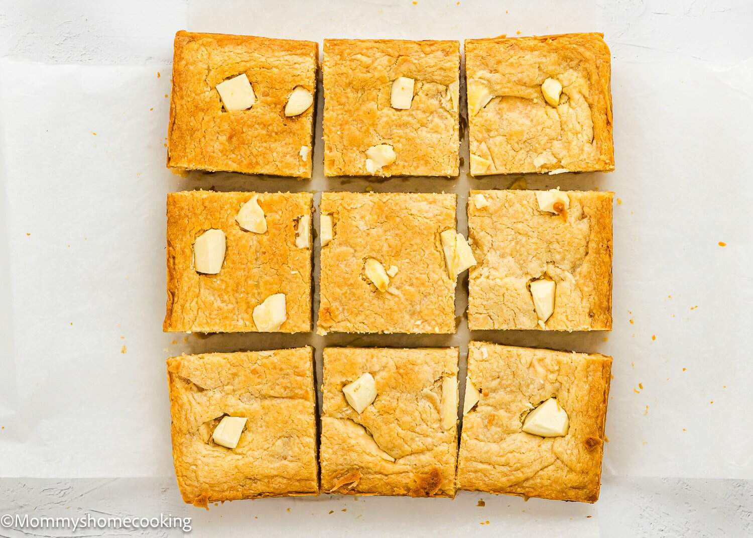 A batch of blondies cut into nine squares, each topped with pieces of white chocolate, placed on white parchment paper.