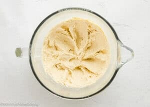 A glass mixing bowl filled with creamy, light-colored dough sits on a white surface.