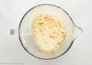 A glass mixing bowl filled with creamy, light tan dough on a white surface.