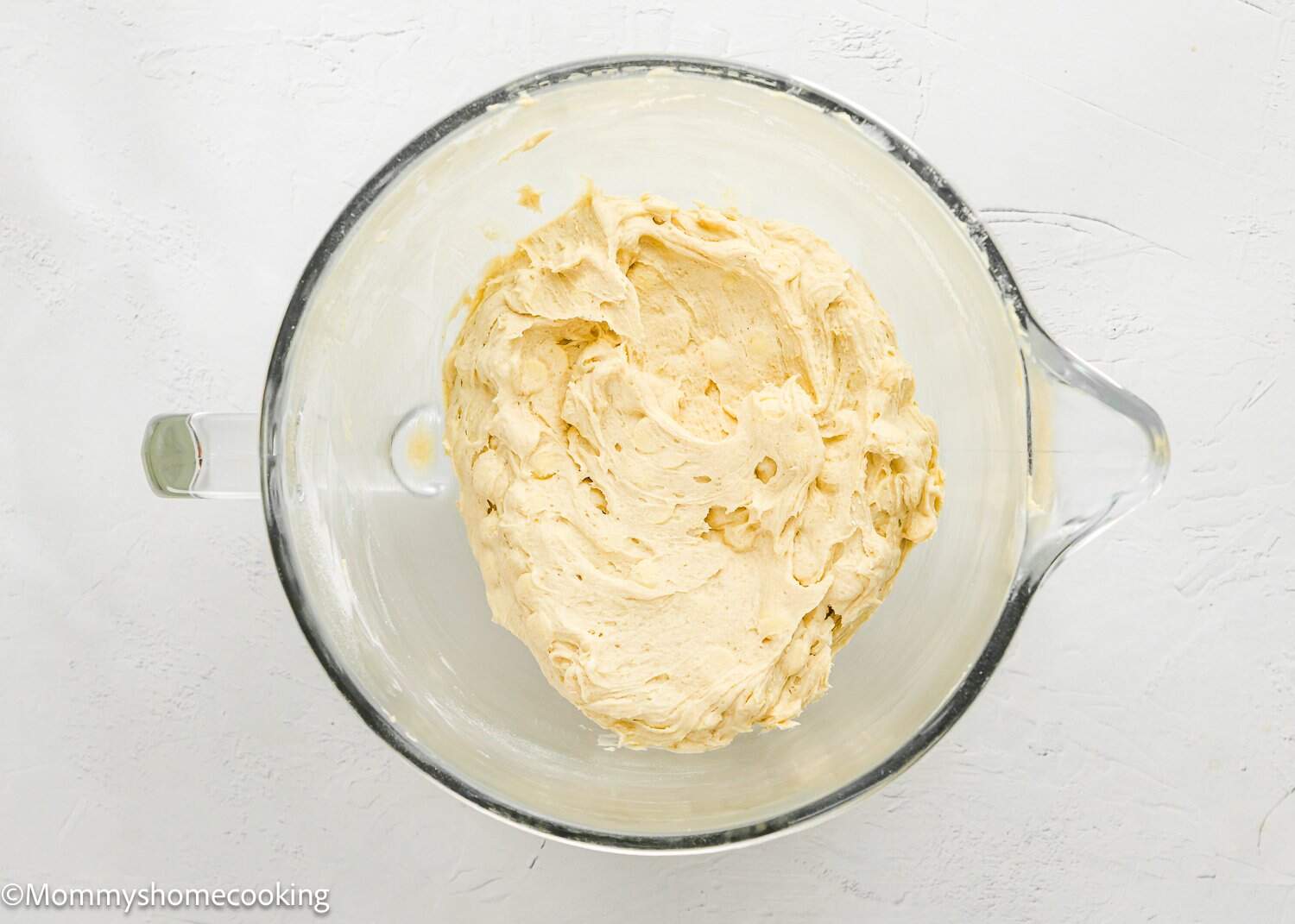 A glass mixing bowl filled with creamy, light tan dough on a white surface.