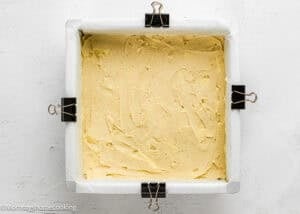 Square baking pan lined with parchment paper and secured with binder clips, filled with a smooth layer of cake batter, ready to be baked.