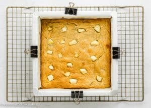 A square baking pan lined with parchment paper holds a golden baked cake with small pieces of butter on top, placed on a cooling rack.