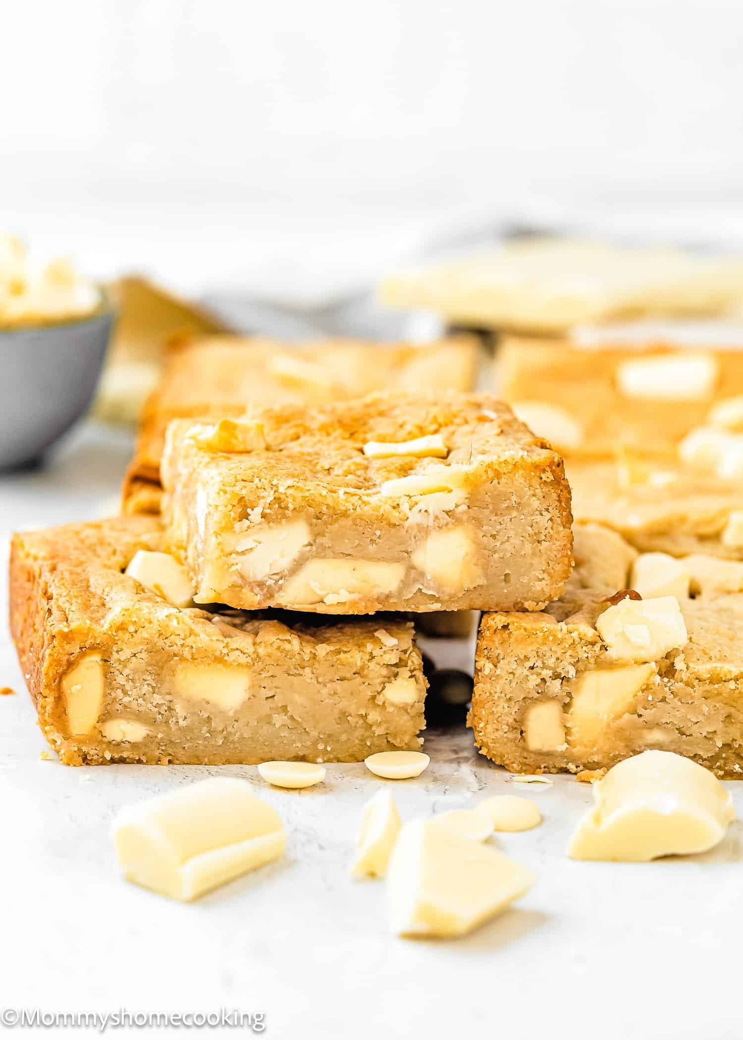 Three eggless white chocolate blondies are stacked in the foreground, with chunks of white chocolate visible inside and around them. More blondies and a bowl of white chocolate chips are in the background.