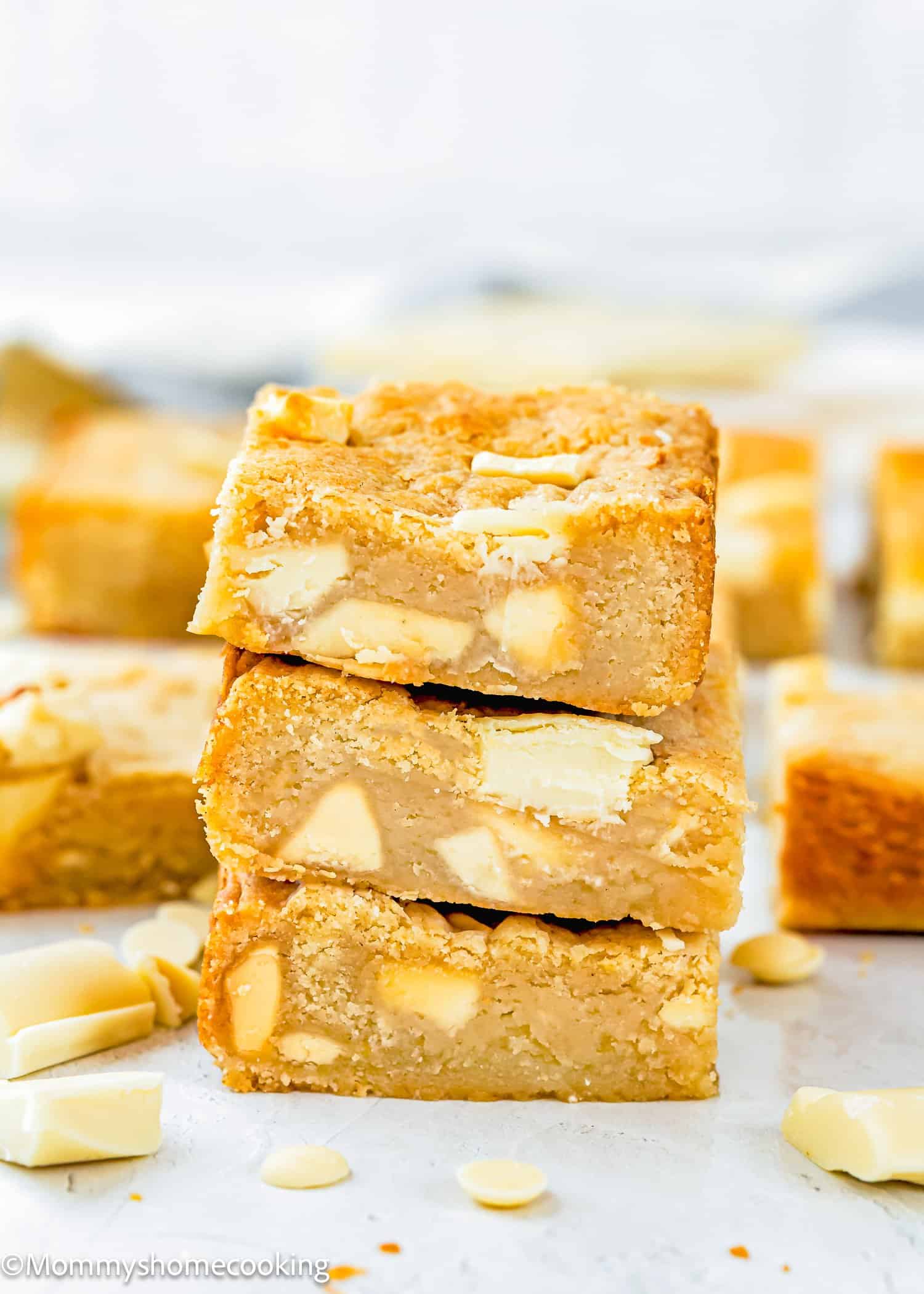 Three stacked white chocolate blondies with visible white chocolate chunks, surrounded by more blondies and chocolate pieces on a light surface.