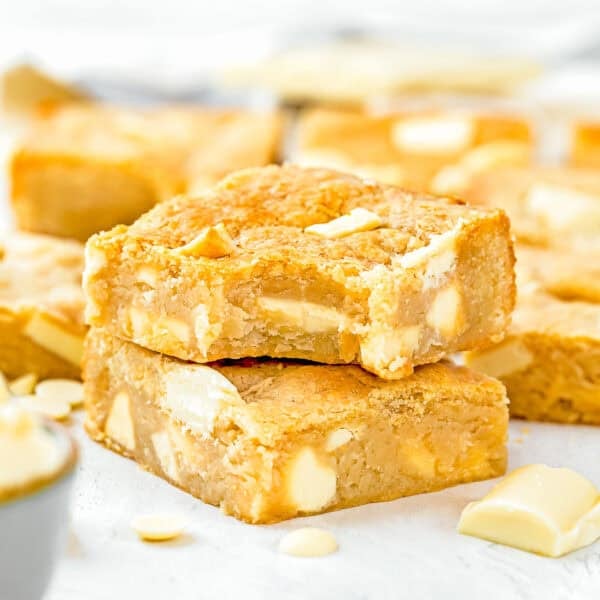 Two egg-free blondie dessert bars with white chocolate chunks stacked on a white surface, surrounded by more blondies and white chocolate pieces in the background.