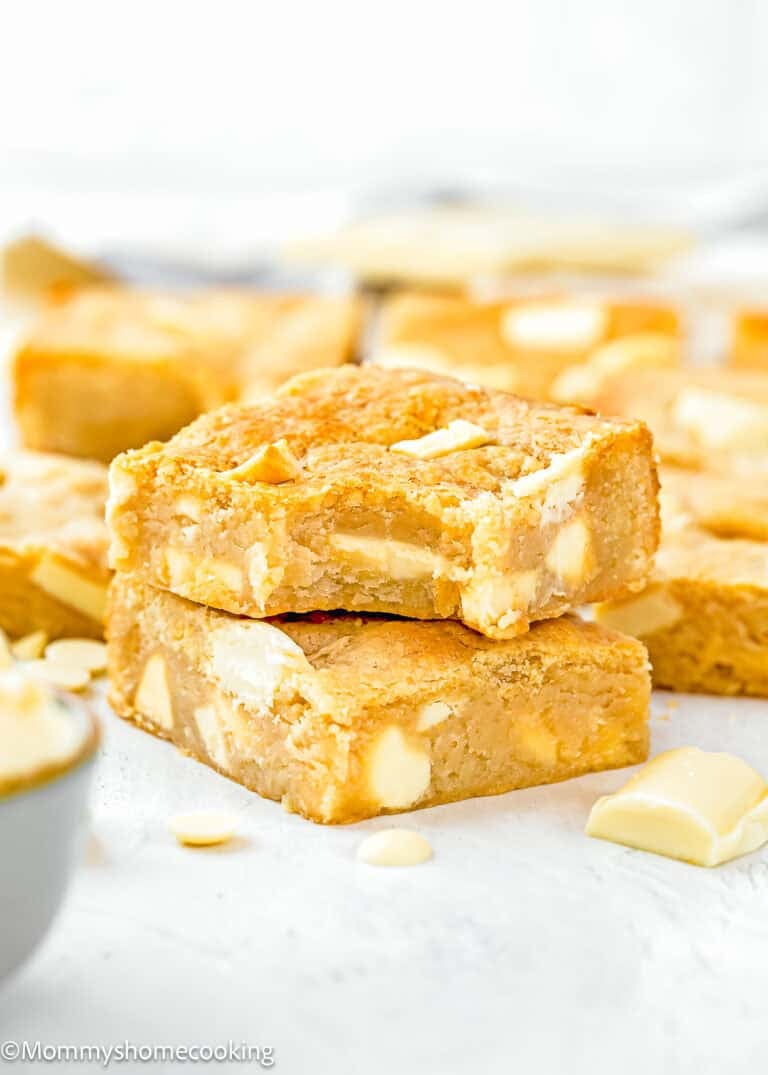 Two egg-free blondie dessert bars with white chocolate chunks stacked on a white surface, surrounded by more blondies and white chocolate pieces in the background.