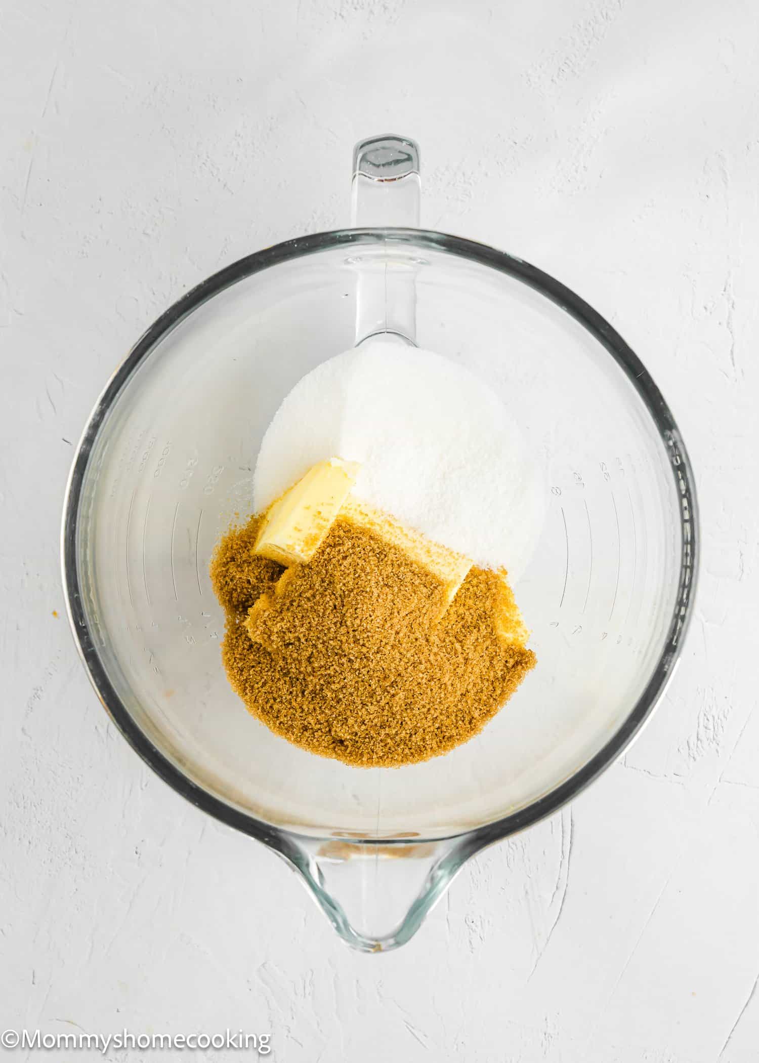 Glass mixing bowl containing granulated sugar, brown sugar, and a stick of butter on a white surface.