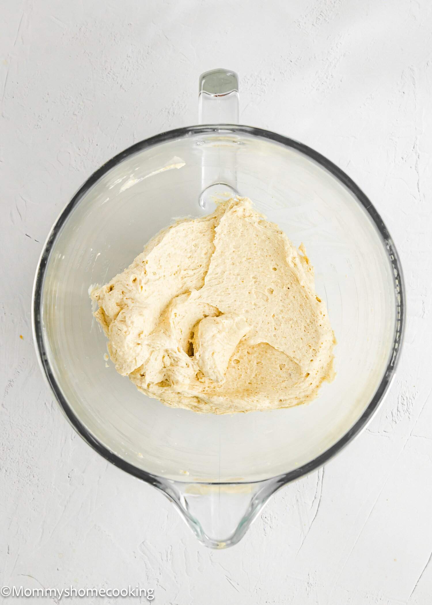 A glass mixing bowl containing partially mixed dough sits on a white textured surface.
