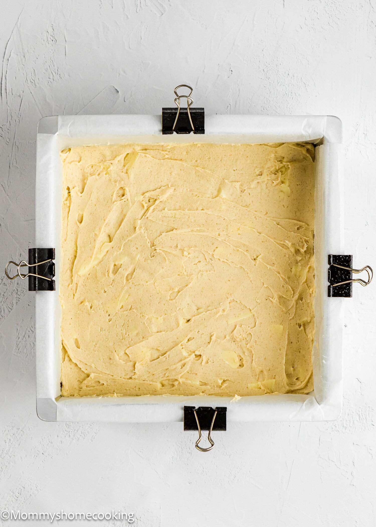 A square baking pan lined with parchment paper and secured with binder clips, filled with uncooked cake batter, on a white surface.