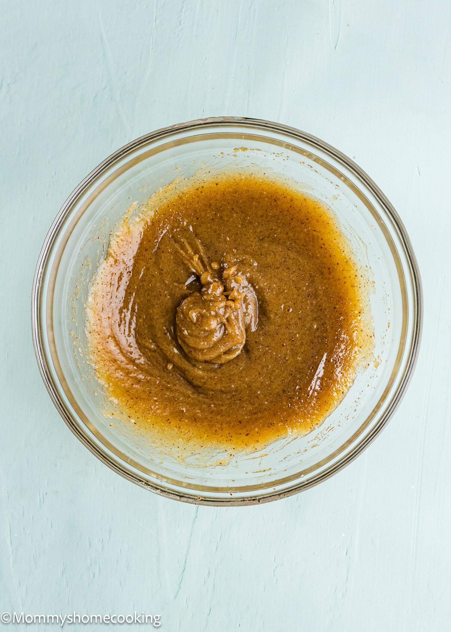 A glass bowl containing a thick, brown batter or mixture sits on a light blue surface.