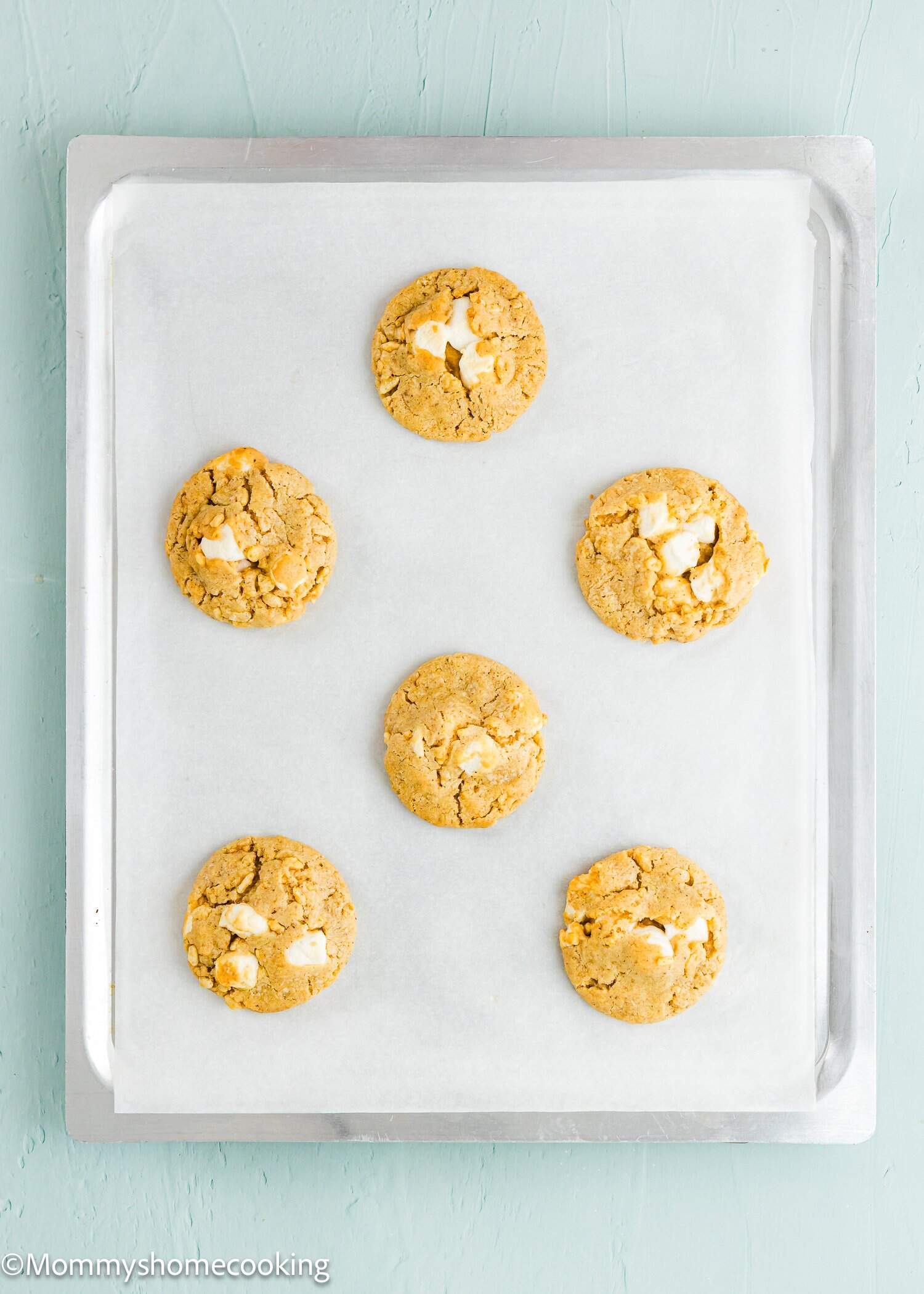 Six baked cookies with visible white chocolate chunks on a parchment-lined baking sheet, viewed from above.