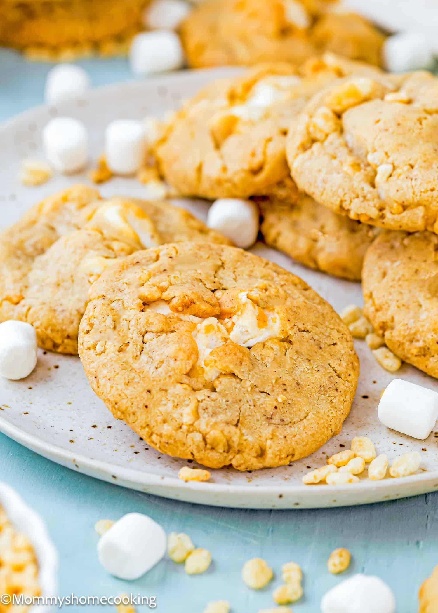 A plate of golden Easy Marshmallow Rice Crispy Cookies (Costco Copycat – Egg-Free) with bits of white marshmallow and crispy rice, surrounded by mini marshmallows and more cookies in the background.