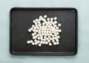A black baking tray with a pile of small white mini marshmallows evenly spread on a light blue surface.