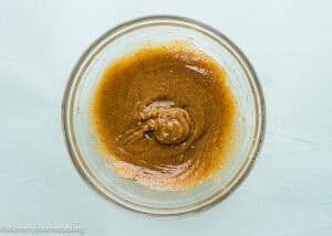 A glass bowl filled with a thick brown mixture, likely a batter or sauce, sits on a light blue surface.