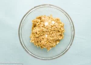 A glass bowl filled with chunky cookie dough mixture set on a light blue surface.