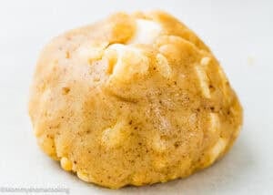 A close-up of a single, round, golden-brown cookie with visible white chocolate chunks on a light background.
