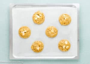 A baking tray lined with parchment paper holds six evenly spaced cookies with white chocolate chunks.