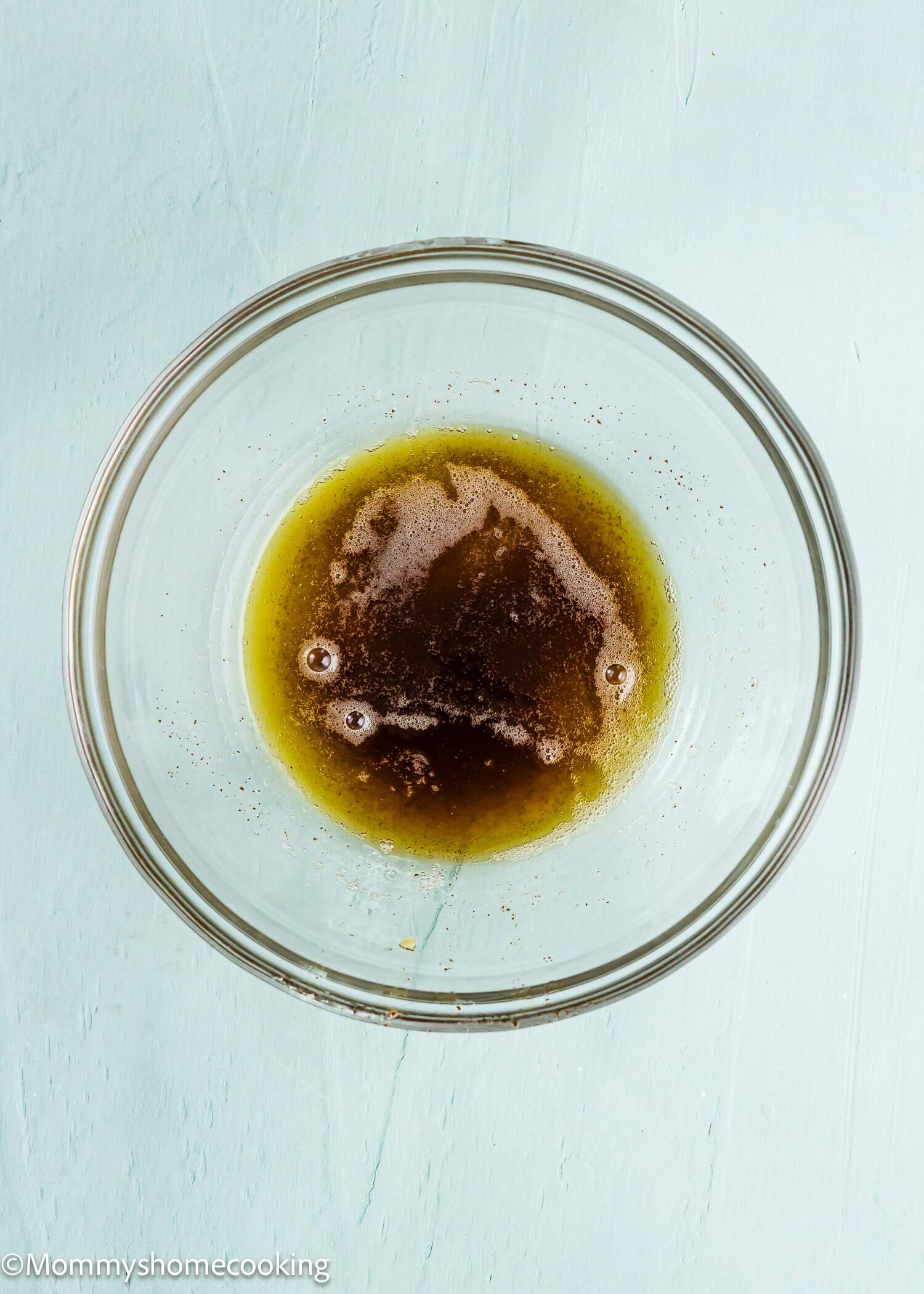 A glass bowl containing melted butter with brown bits, placed on a light blue surface.