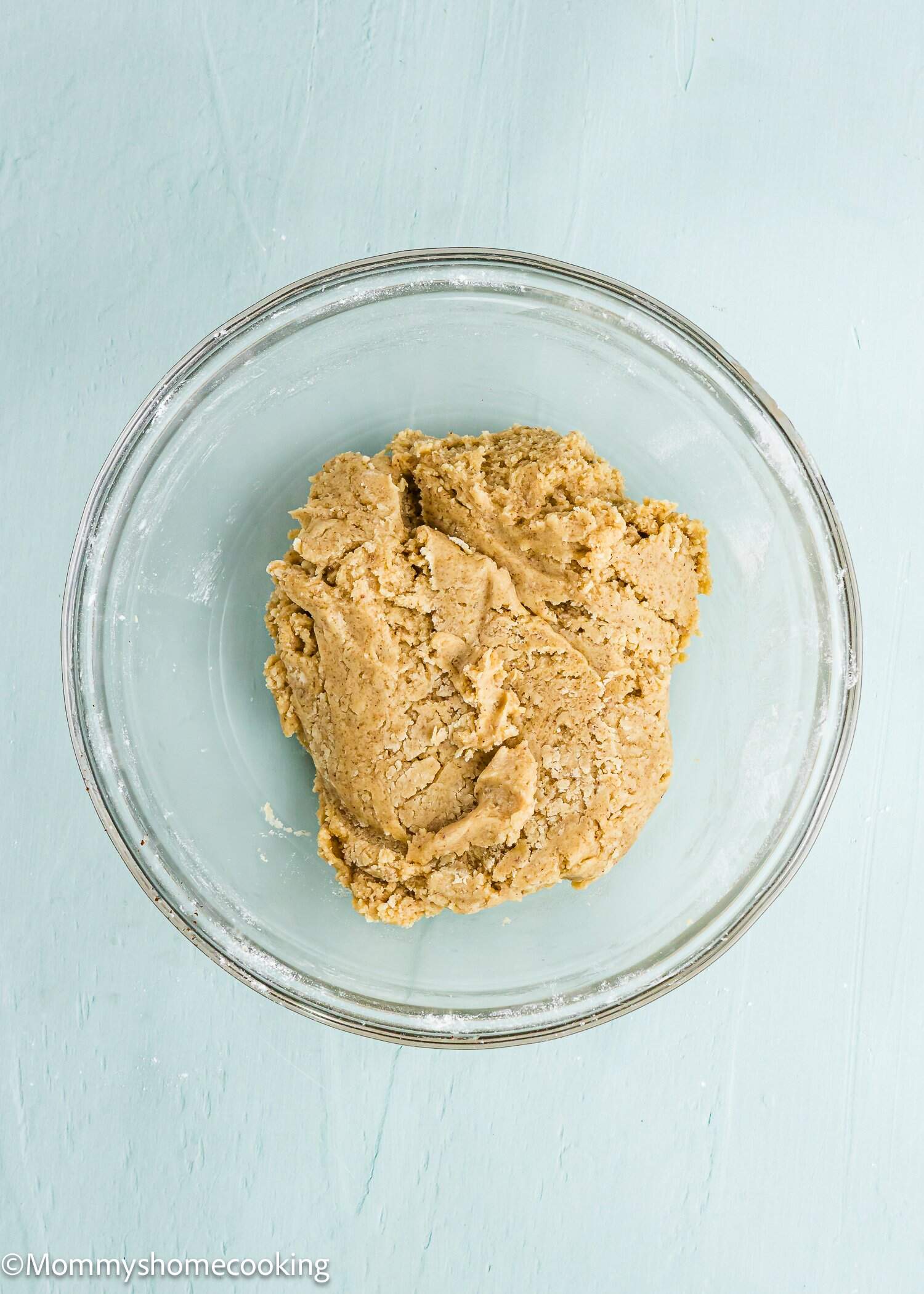 A glass bowl containing a mound of raw cookie dough on a light blue surface.
