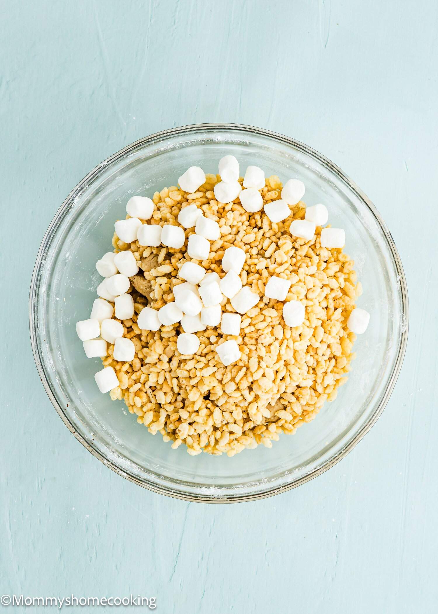A glass bowl filled with rice cereal and mini marshmallows on a light blue surface.