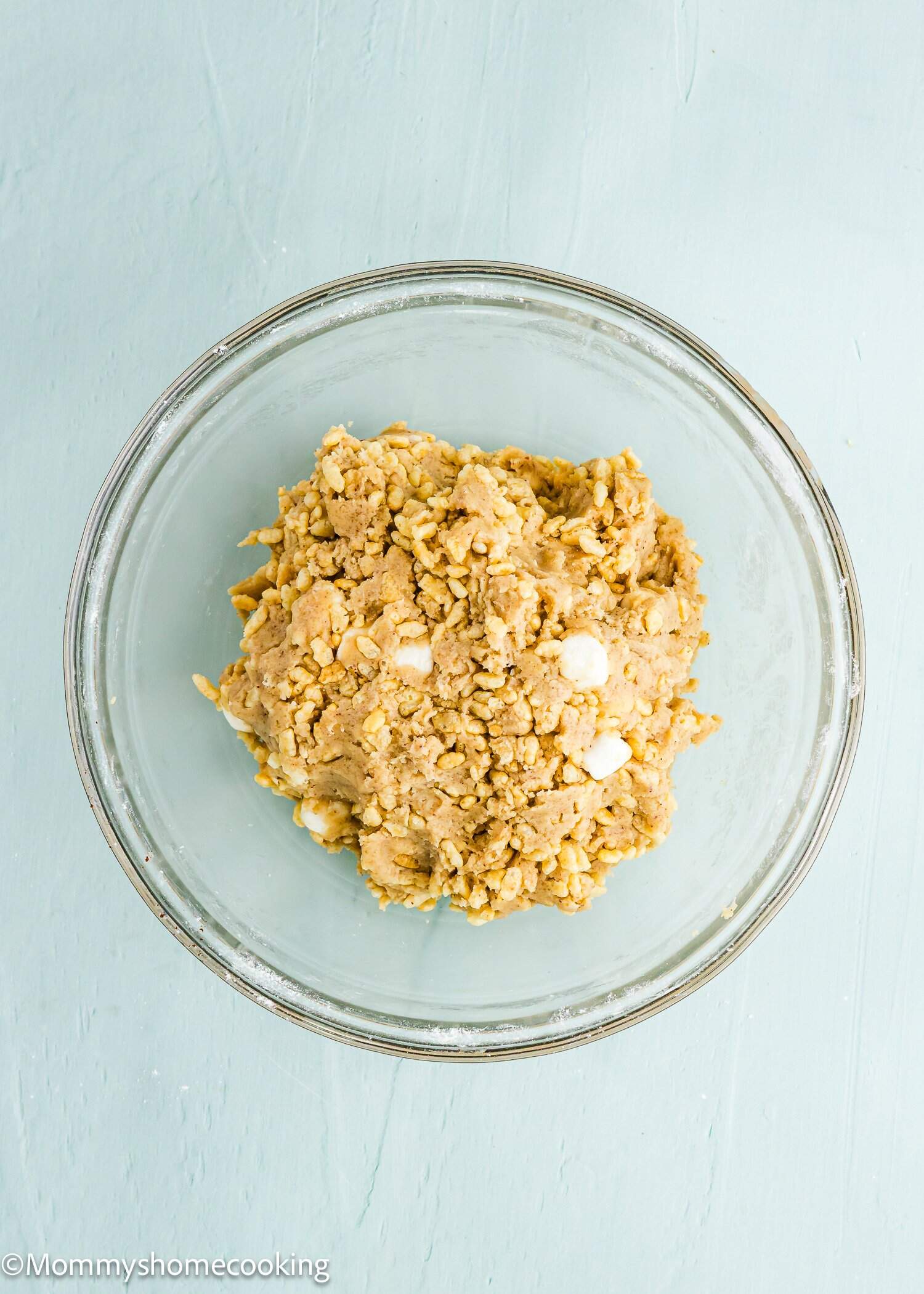A glass bowl containing a mixture of uncooked cookie dough with visible white chocolate chips on a light blue surface.