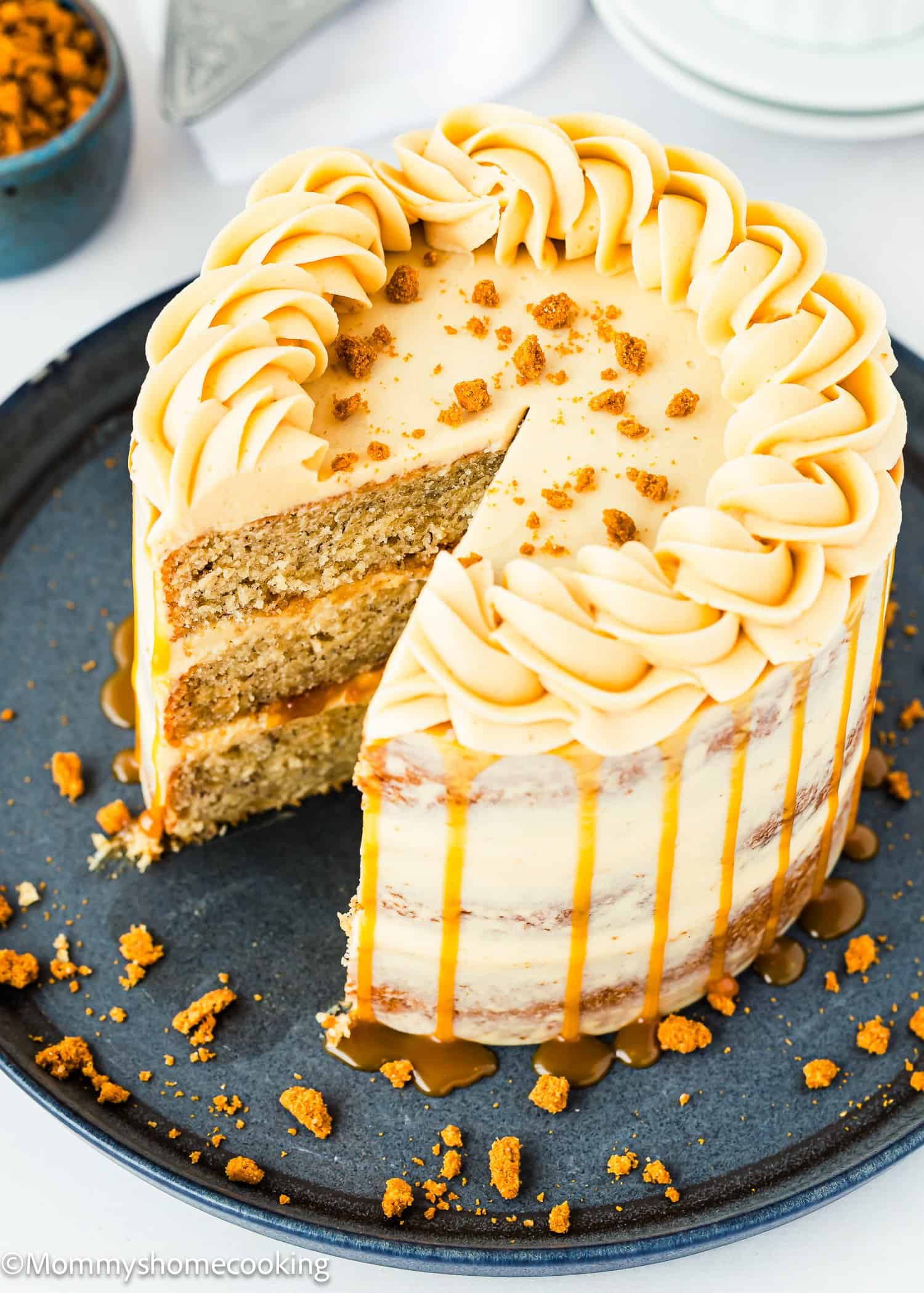 A round, frosted cake with caramel drizzle and cookie crumbs on top sits on a dark plate. A single slice is cut and pulled outward, showing moist cake layers and creamy frosting. Crumbs and caramel surround the cake on the plate.