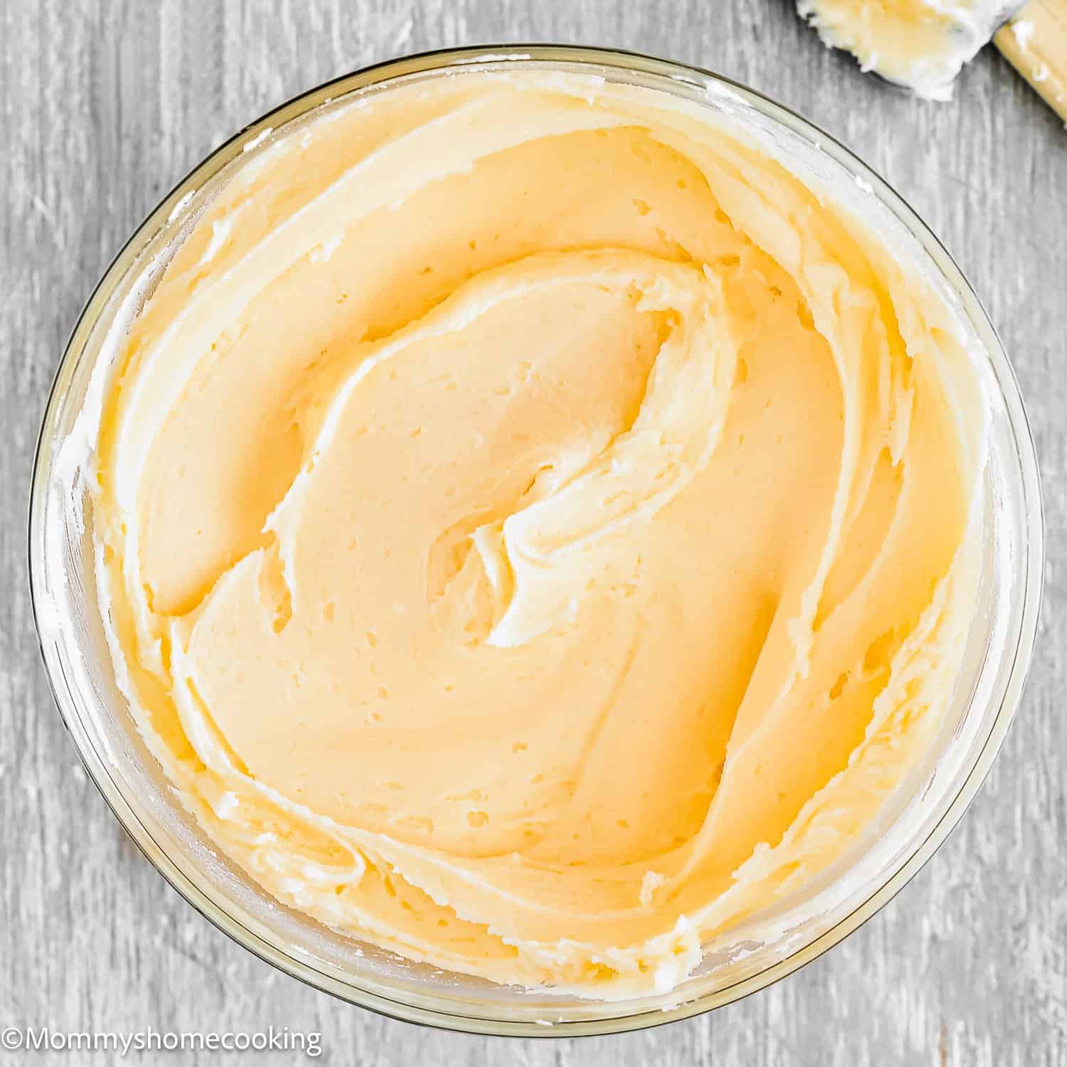 A glass bowl filled with smooth, creamy caramel buttercream frosting sits on a light gray wooden surface. A small amount of frosting is on a spatula in the upper right corner.
