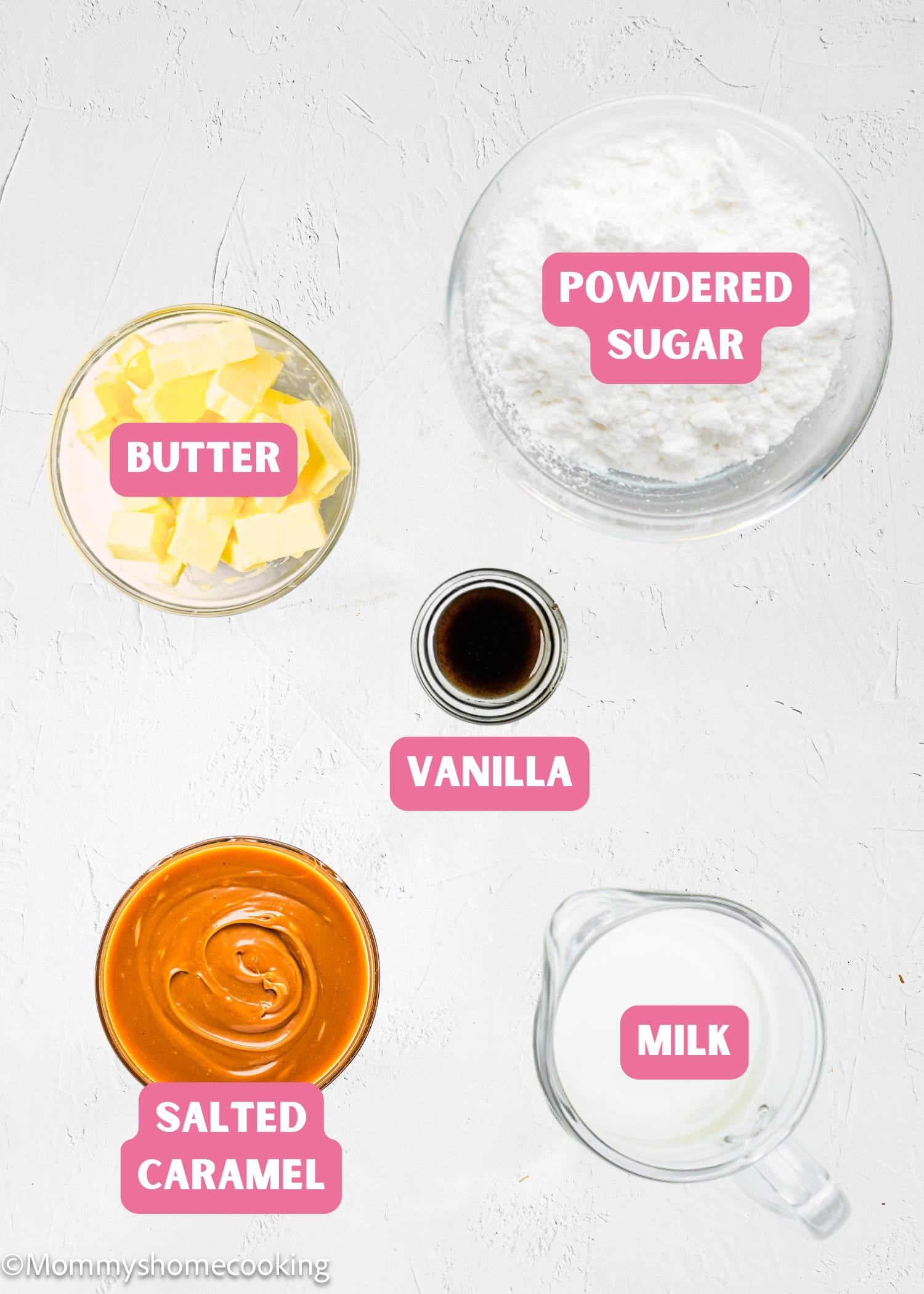 Five labeled ingredients for a recipe arranged on a white surface: powdered sugar, butter, vanilla, salted caramel, and milk, each in a separate glass container.