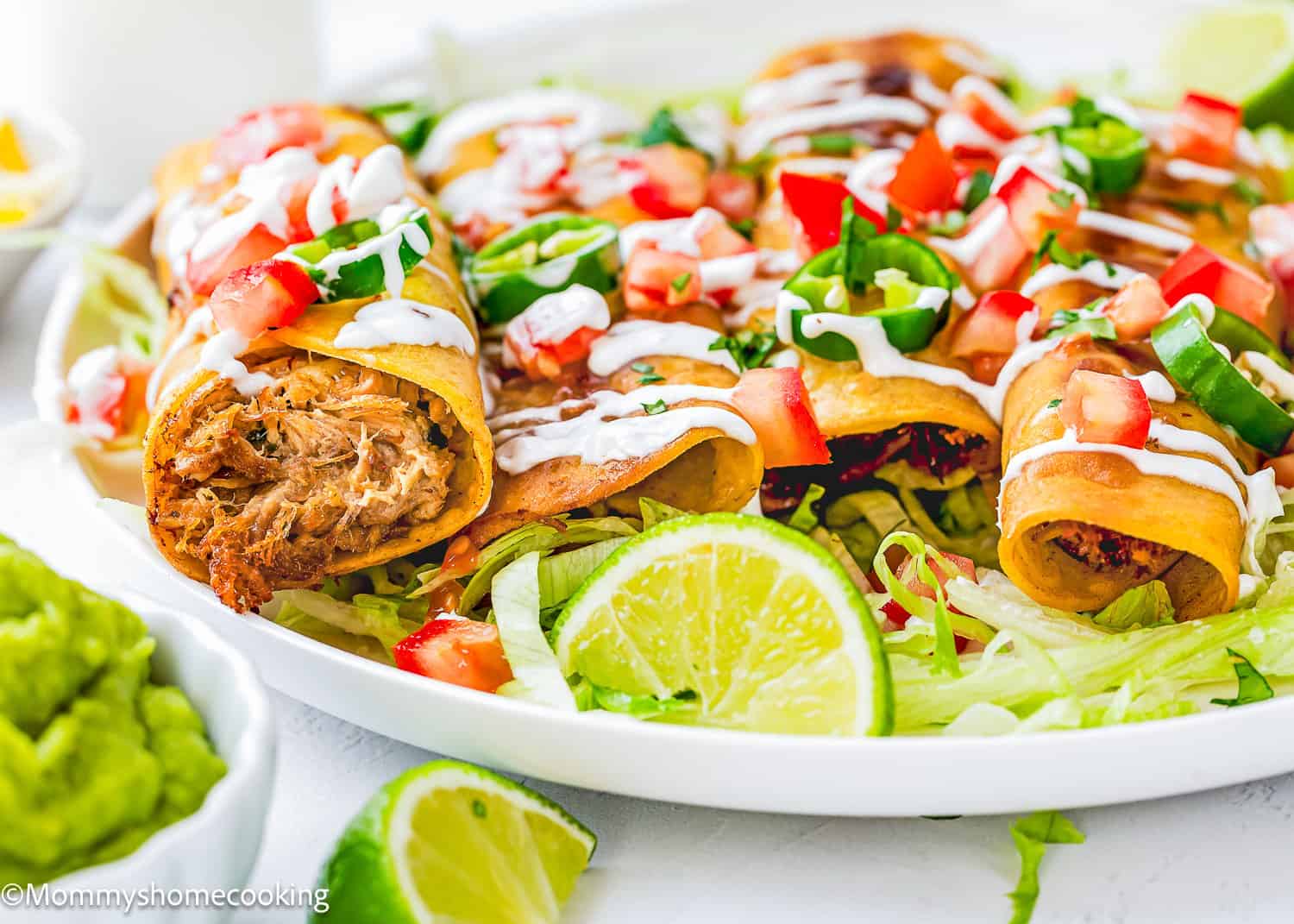 A plate of crispy taquitos filled with shredded meat, topped with diced tomatoes, jalapenos, and drizzled with sour cream, served on a bed of shredded lettuce with lime wedges and guacamole on the side.