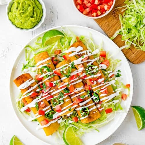 A plate of caritas taquitos topped with chopped tomatoes, shredded lettuce, and drizzled with crema. Sides of guacamole, shredded cheese, lettuce, and lime wedges surround the dish on a white surface.