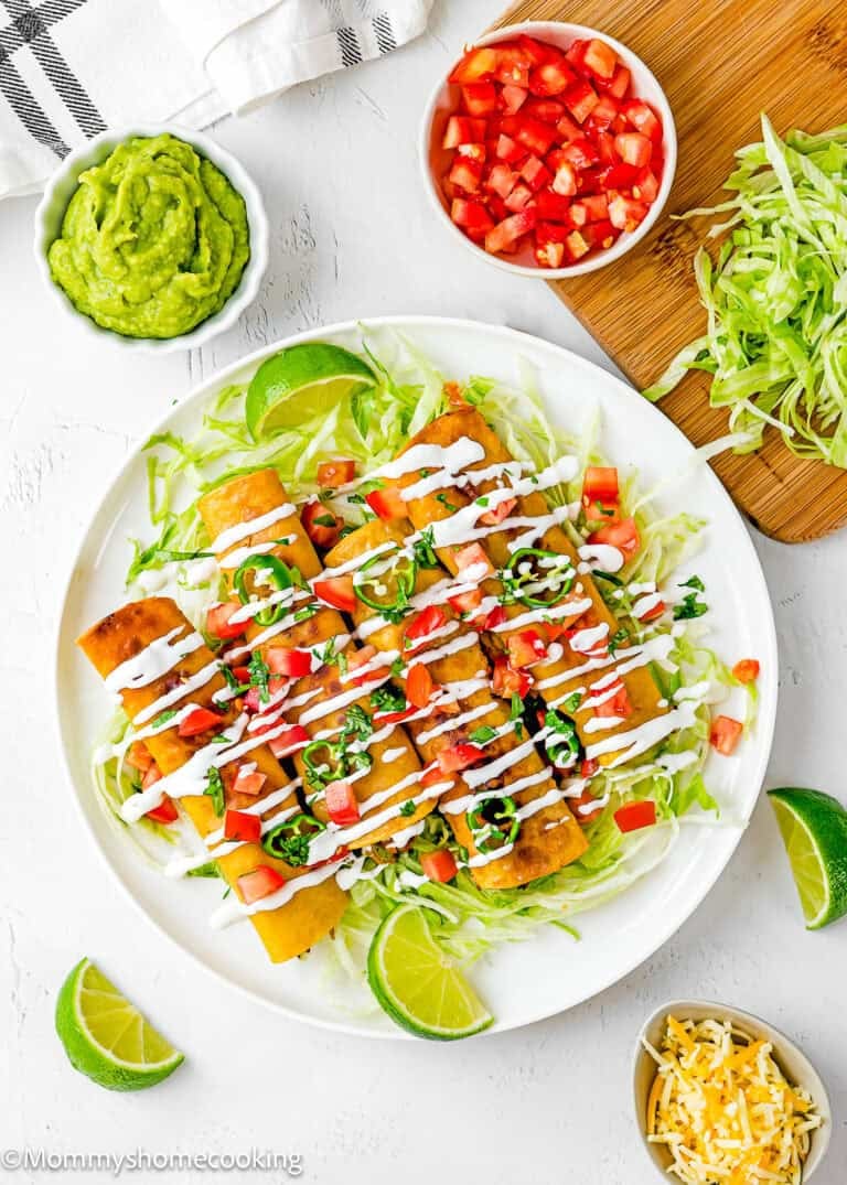 A plate of caritas taquitos topped with chopped tomatoes, shredded lettuce, and drizzled with crema. Sides of guacamole, shredded cheese, lettuce, and lime wedges surround the dish on a white surface.