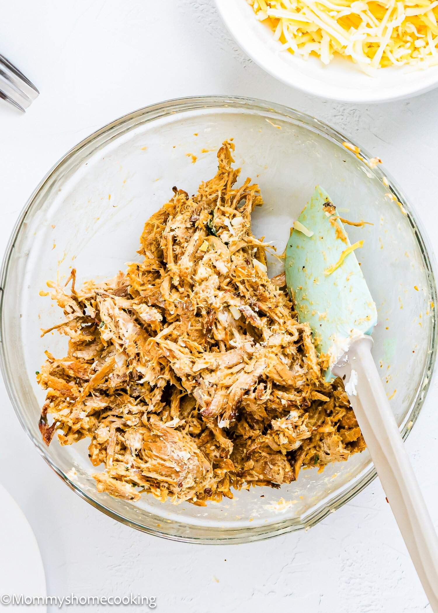 A glass bowl filled with shredded, seasoned carnitas and a spatula, placed on a white surface. Shredded cheese is visible in a separate bowl nearby.