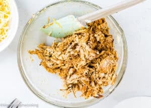A glass bowl filled with shredded seasoned meat being mixed with a spatula, set on a light-colored surface. A bowl with shredded cheese is partially visible to the side.