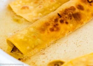 Close-up of golden-brown, crispy taquitos or rolled tortillas in a shallow layer of oil, highlighting their texture and fried surface.