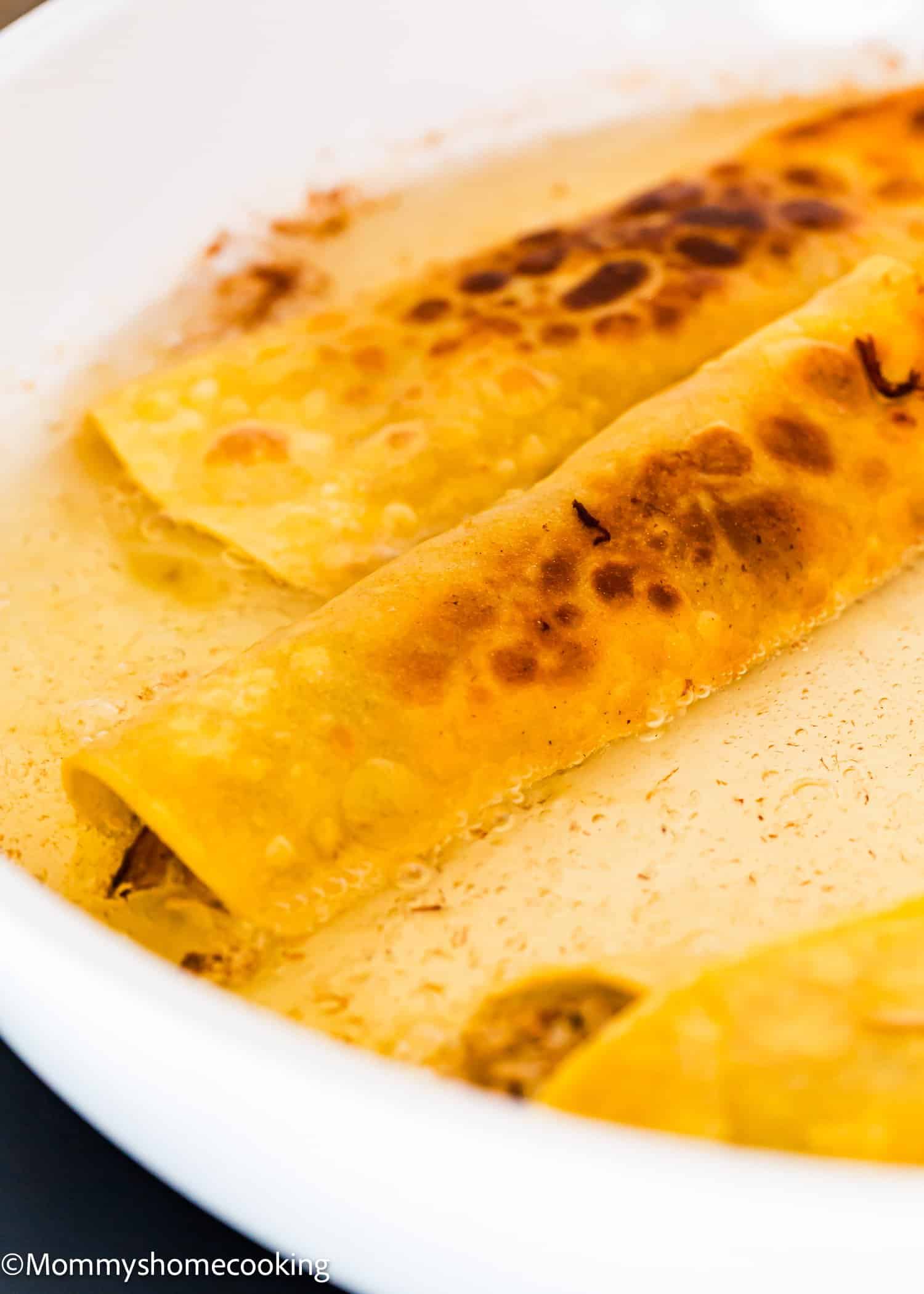Two golden-brown rolled tortillas sit in a white dish with a creamy sauce, showing toasted spots on the surface. The image is close-up and highlights the texture of the tortillas.