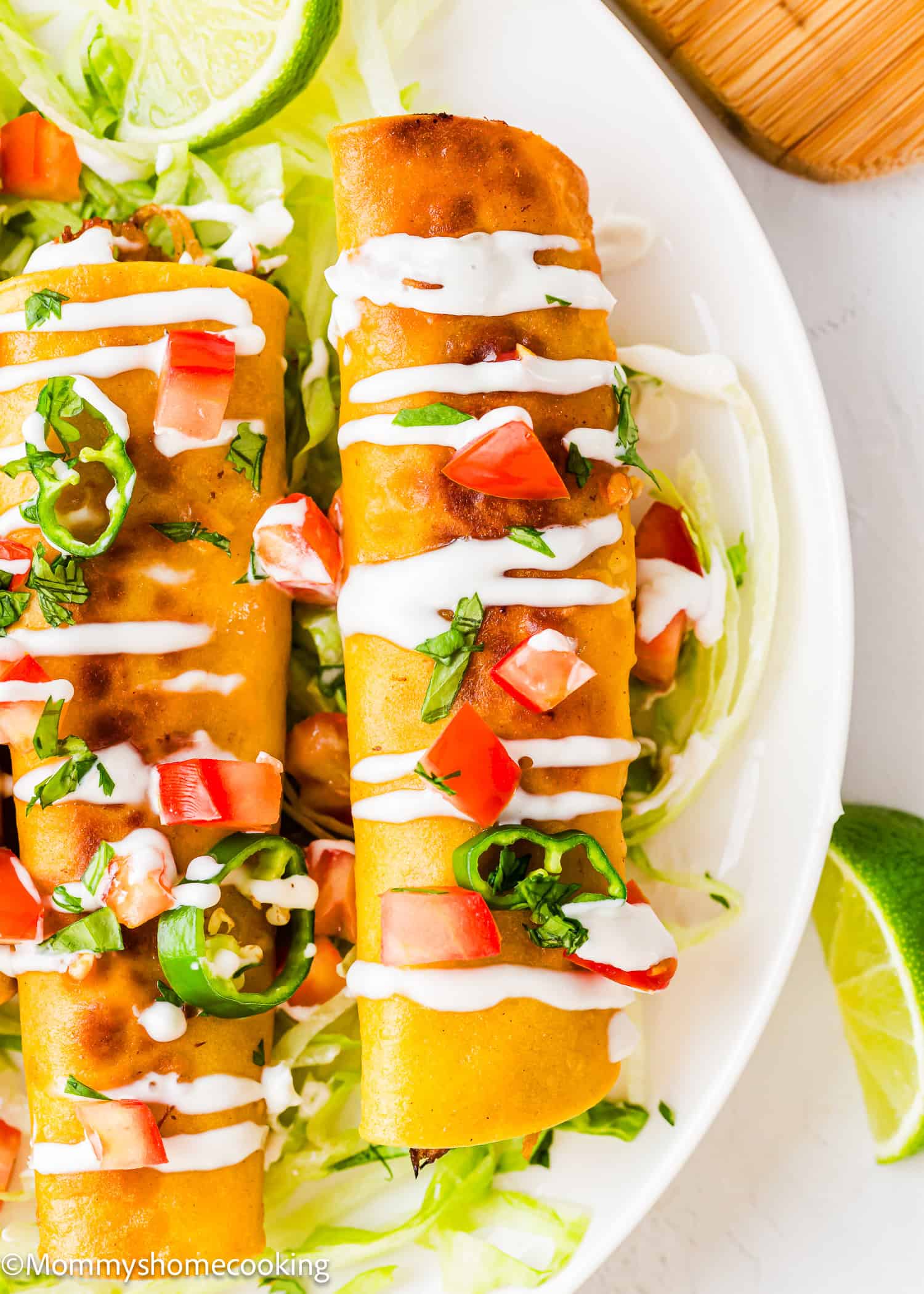 Two golden, crispy taquitos topped with chopped tomatoes, jalapeños, fresh herbs, and drizzled with white sauce, served on a bed of shredded lettuce with lime wedges on the side.
