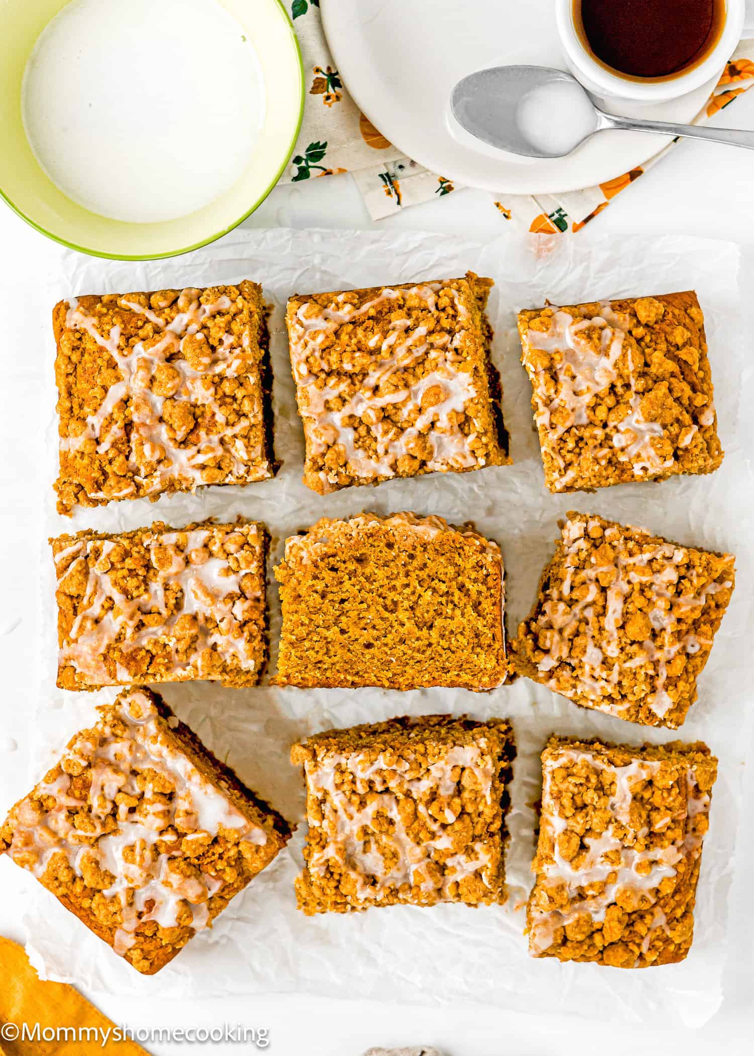 Nine pieces of Egg-Free Pumpkin Coffee Cake with Crumb Topping and icing drizzle are arranged on parchment paper, with a bowl of milk and a cup of coffee nearby.