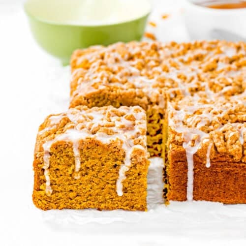 A square Egg-Free Pumpkin Coffee Cake with crumb topping and white icing, sliced to reveal its moist interior. A green bowl and a cup are in the background.