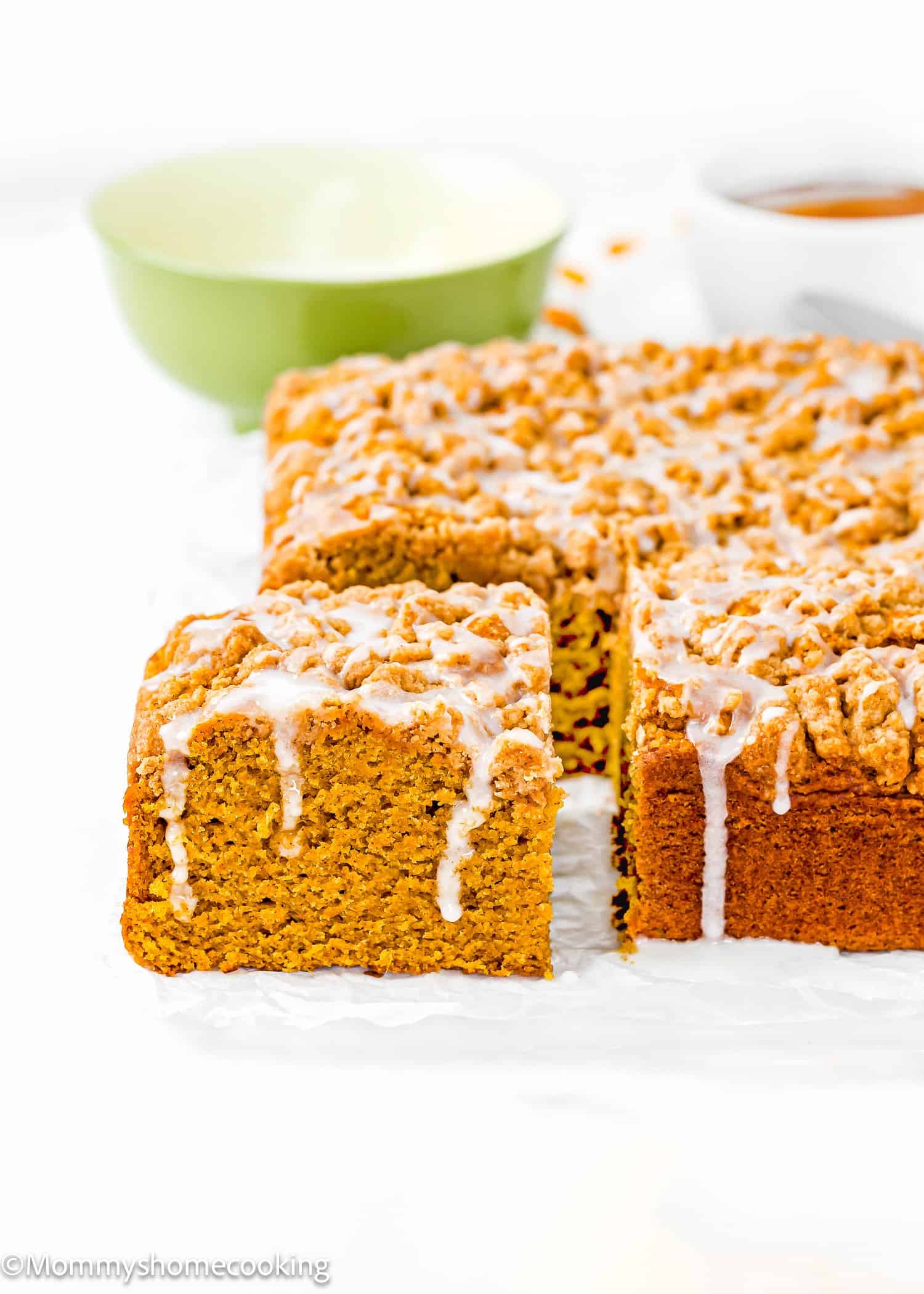 A square Egg-Free Pumpkin Coffee Cake with crumb topping and white icing, sliced to reveal its moist interior. A green bowl and a cup are in the background.