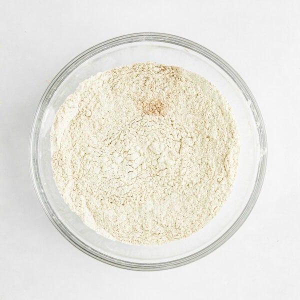 A clear glass bowl filled with dry flour on a white surface.