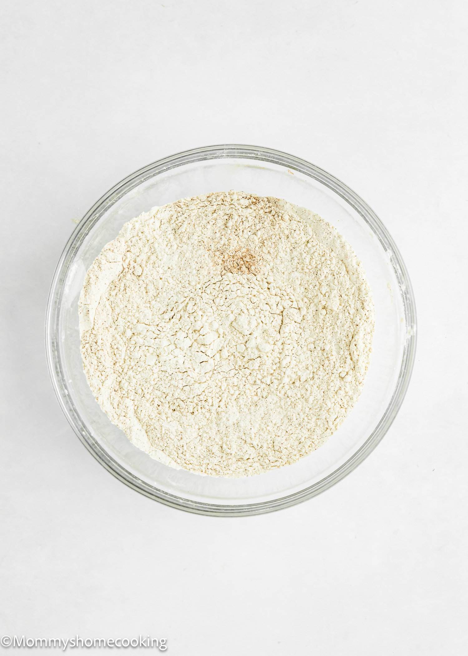 A clear glass bowl filled with dry flour on a white surface.