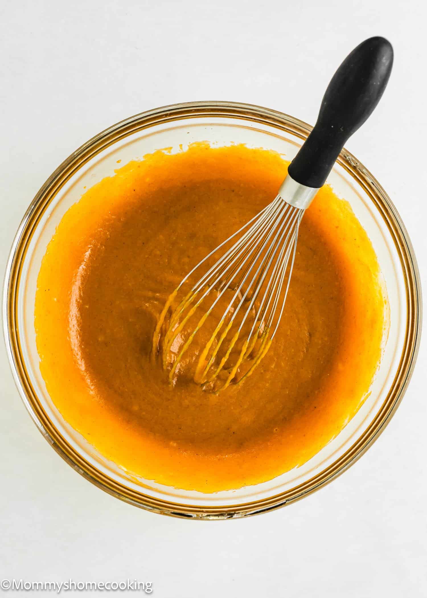 A glass bowl filled with orange batter being mixed with a metal whisk that has a black handle, on a white surface.