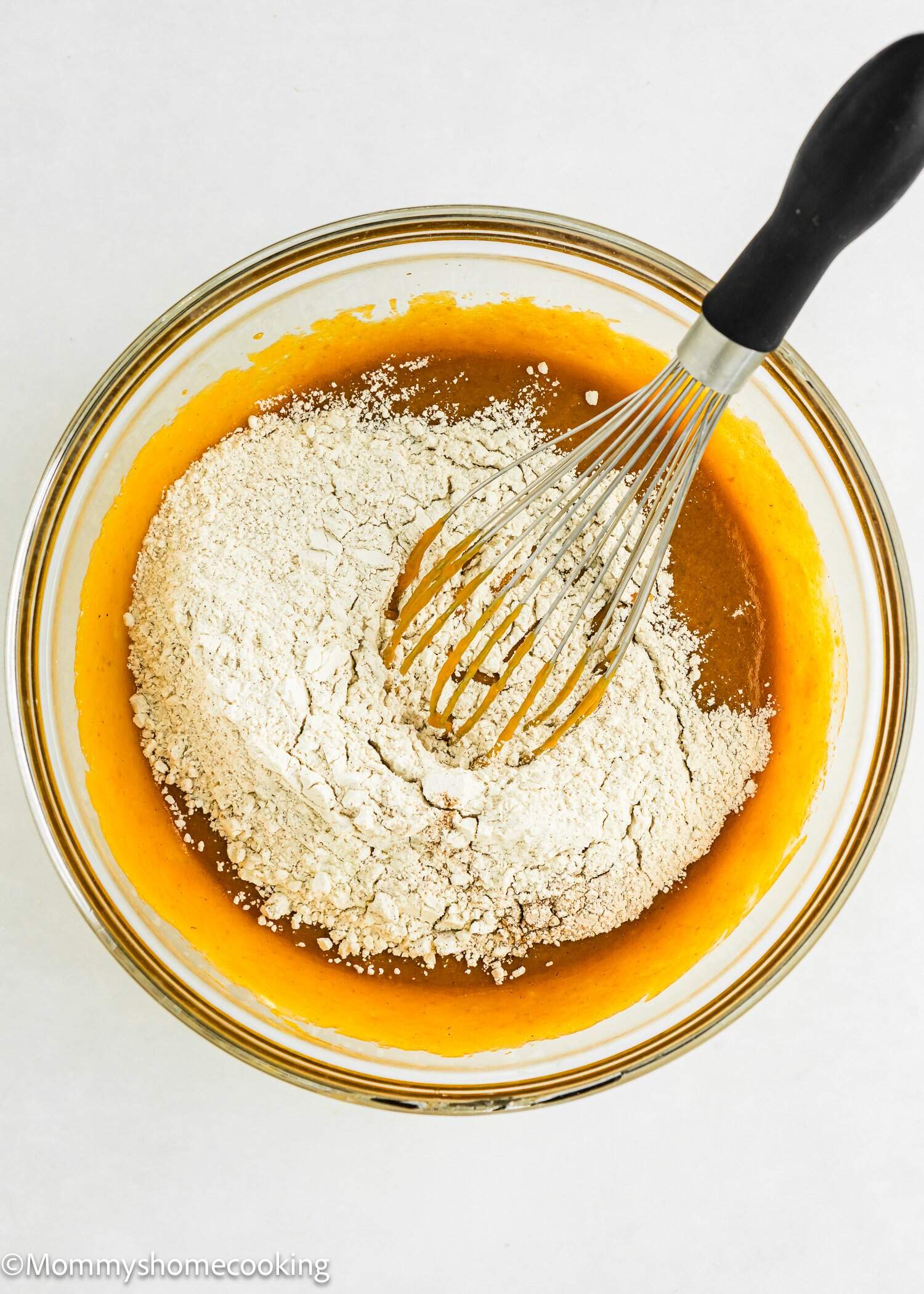 A glass bowl with flour and orange liquid mixture being whisked together on a white surface.