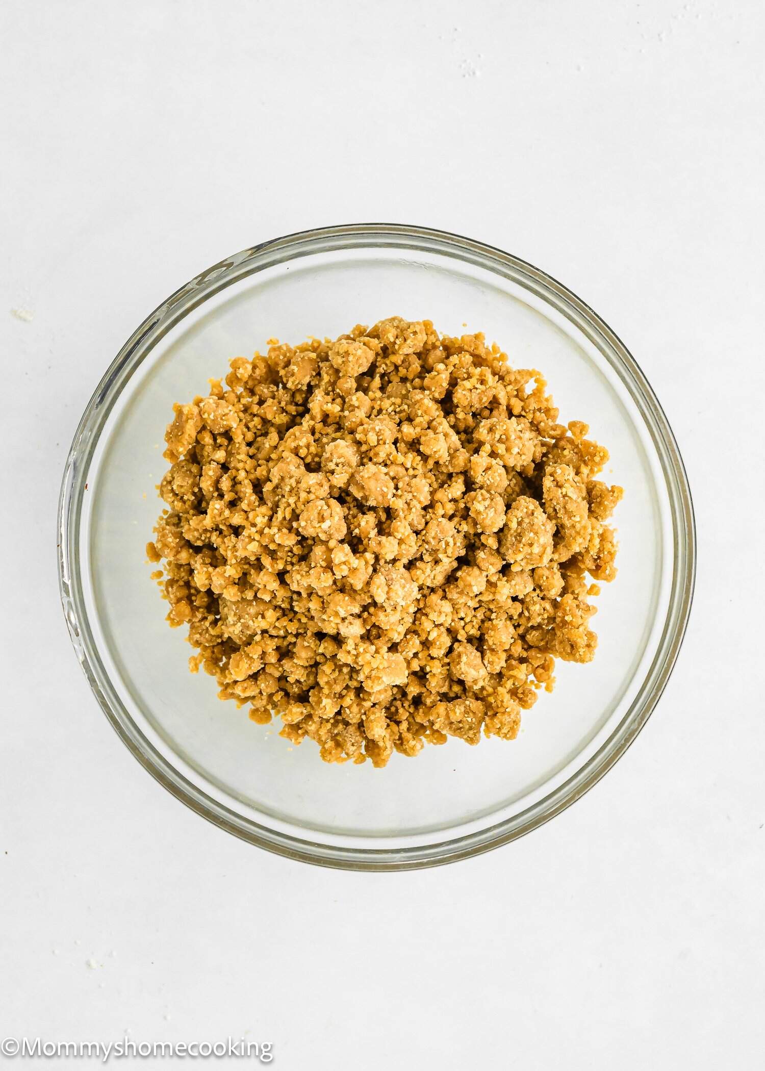 A clear glass bowl filled with crumbly, light brown streusel topping on a white background.
