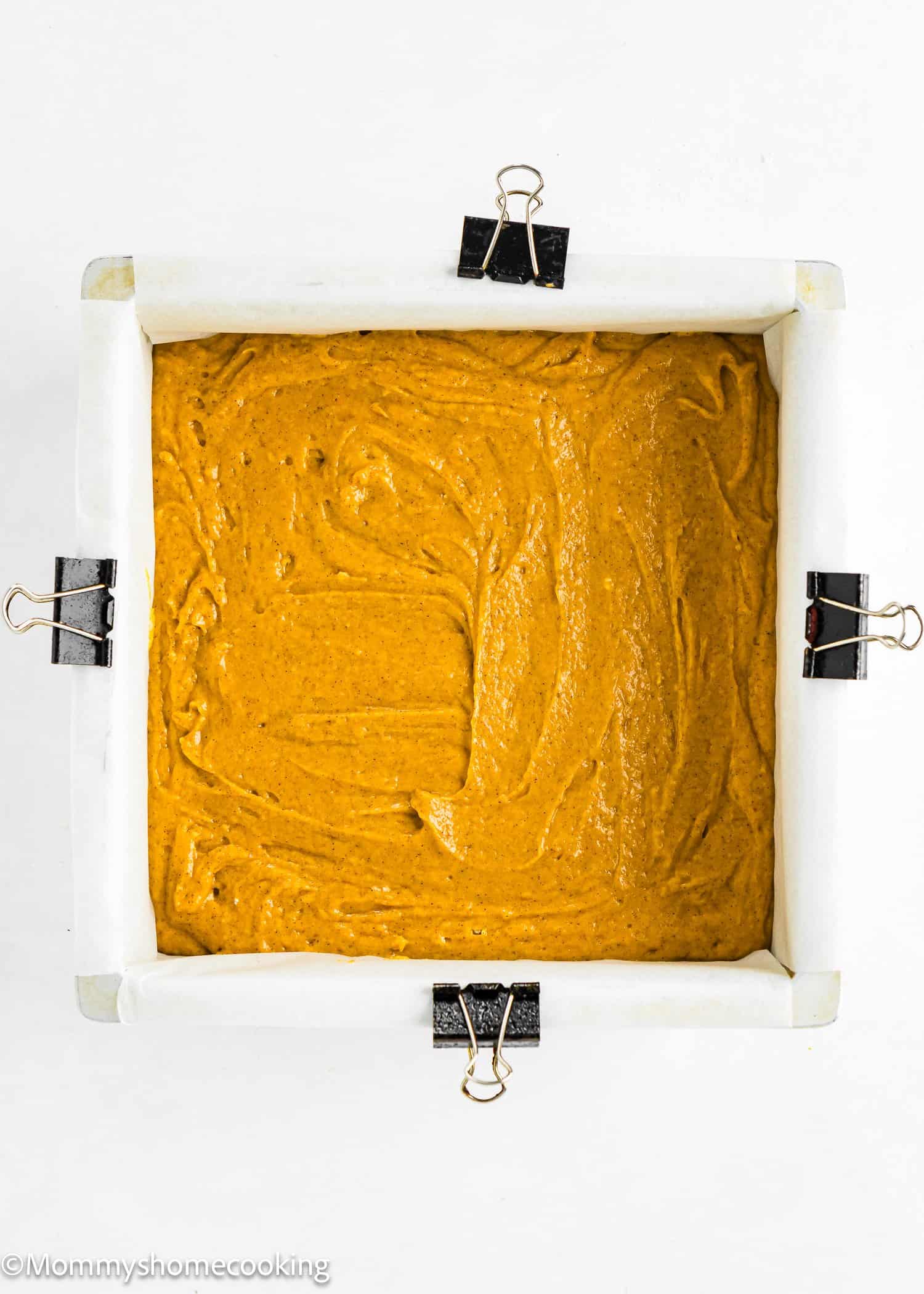 A square baking pan lined with parchment paper and clips, filled with an even layer of uncooked orange-brown batter.
