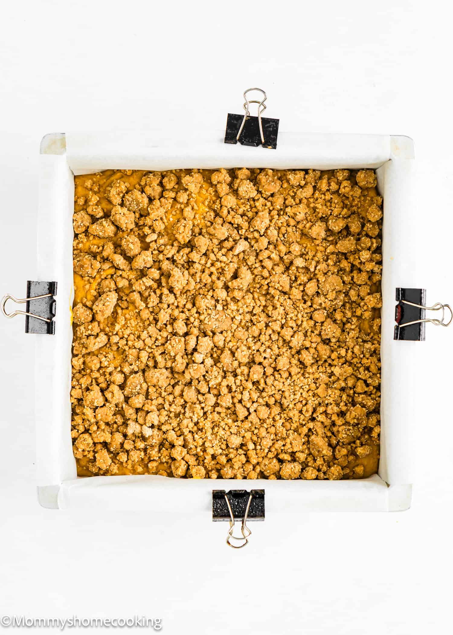 A square pan lined with parchment paper and secured with binder clips, filled with an even layer of crumbly topping.