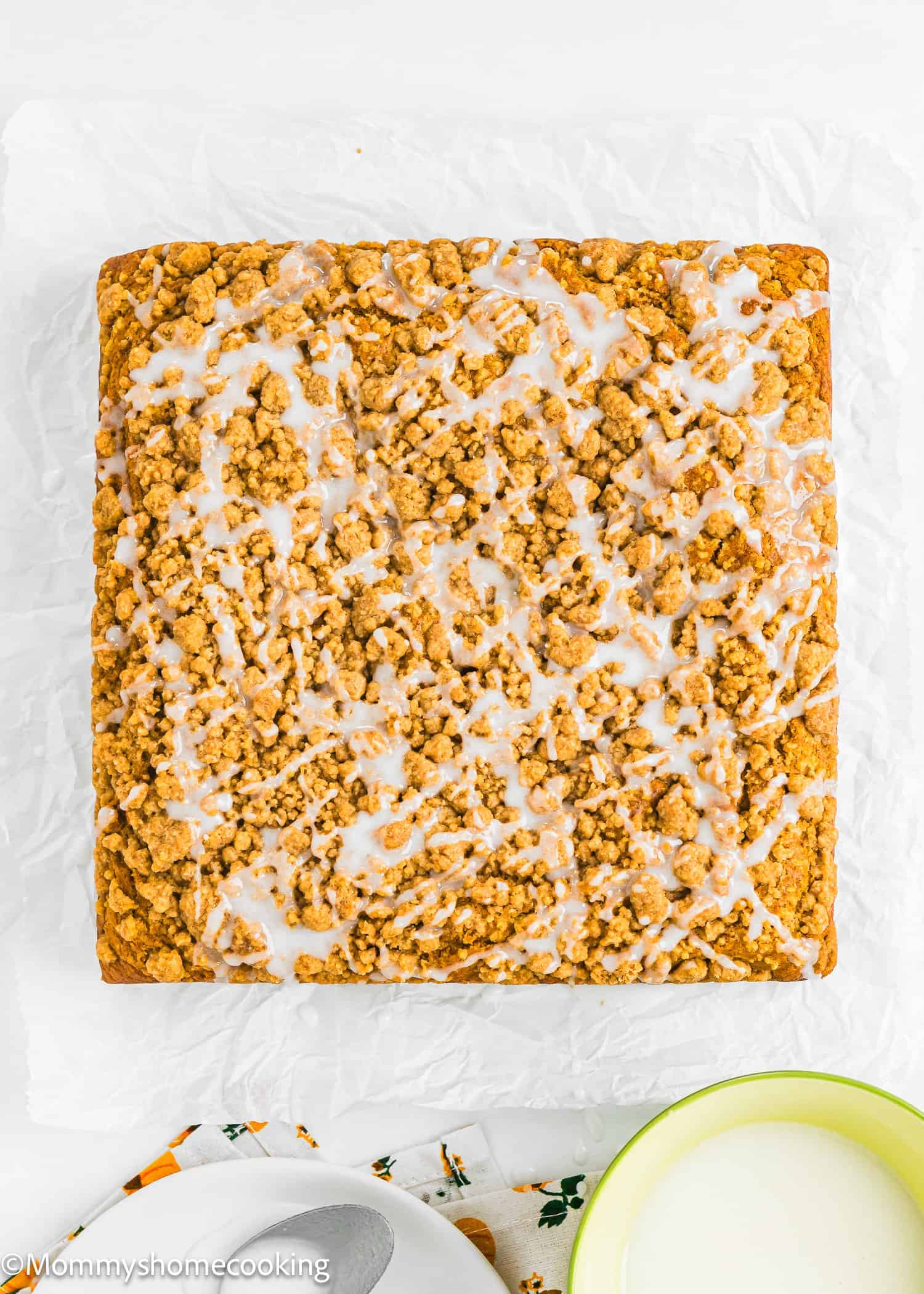 A square crumb cake with icing drizzle on top sits on white parchment paper, next to a bowl of milk and a spoon.