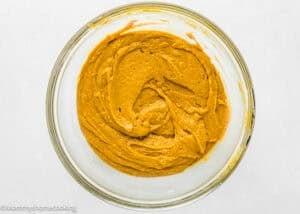 A glass bowl filled with smooth, orange-brown Egg-Free Pumpkin Coffee Cake batter with crumb topping sits on a white surface.