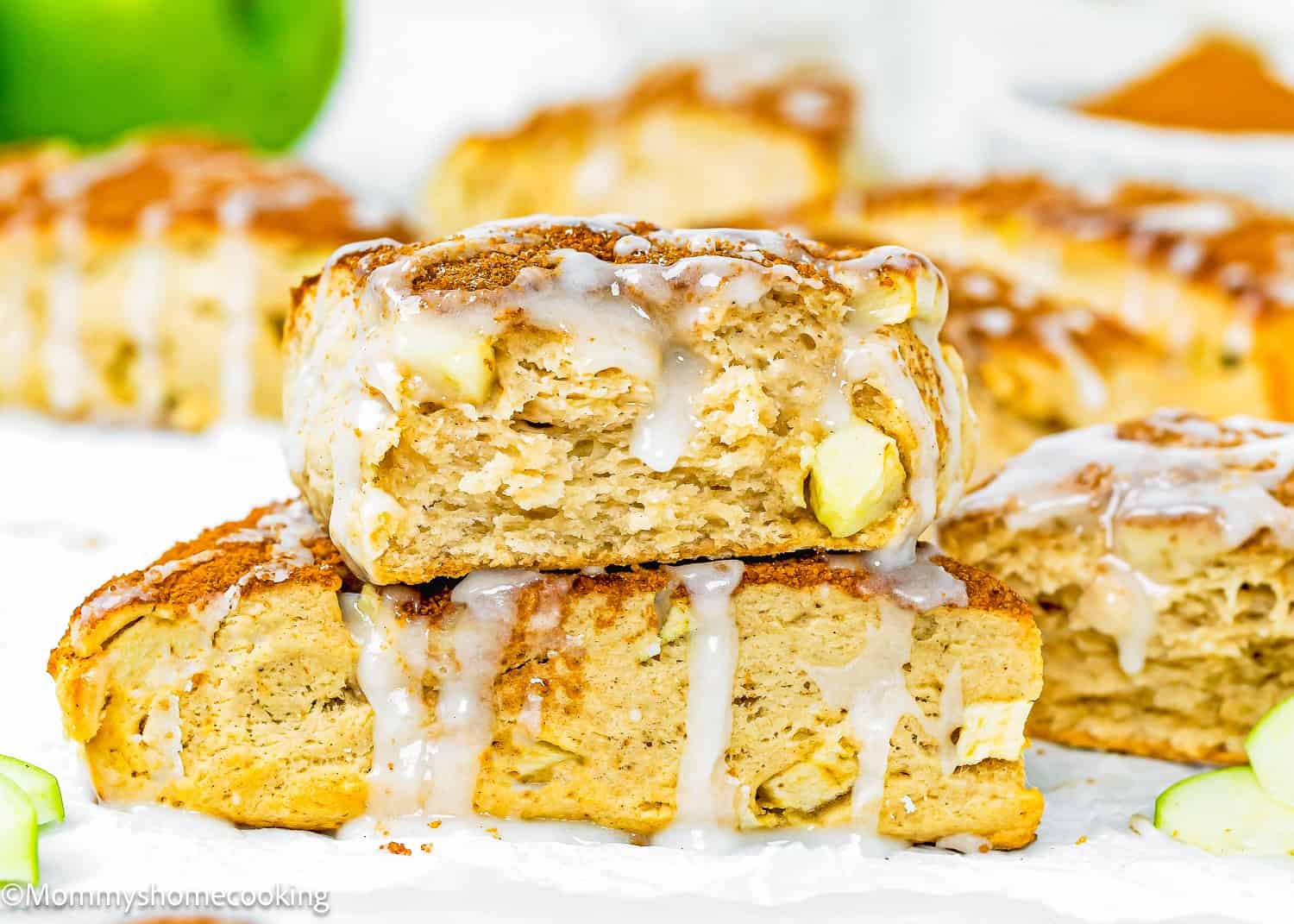 Two eggless apple cinnamon scones stacked on top of each other, topped with a drizzle of white icing, with more scones blurred in the background.